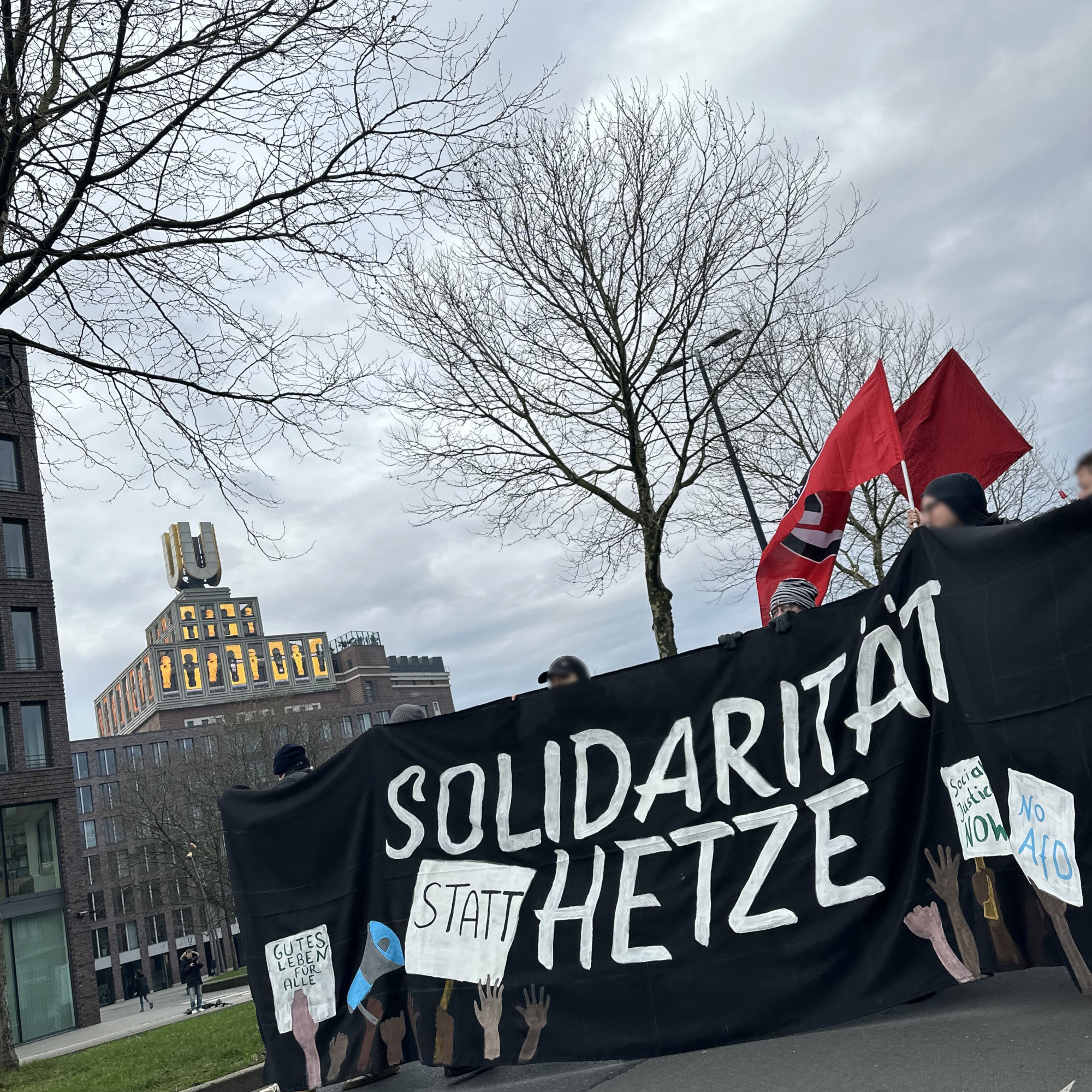 Das Fronttransparent mit der Aufschrift Solidarität statt Hetze, dahinter weht eine Antifafahne und eine rote Fahne. Im Hintergrund das Wahrzeichen Dortmunder U mit einer Videoinstallation mit schwarzen und gelben Kickerfiguren.